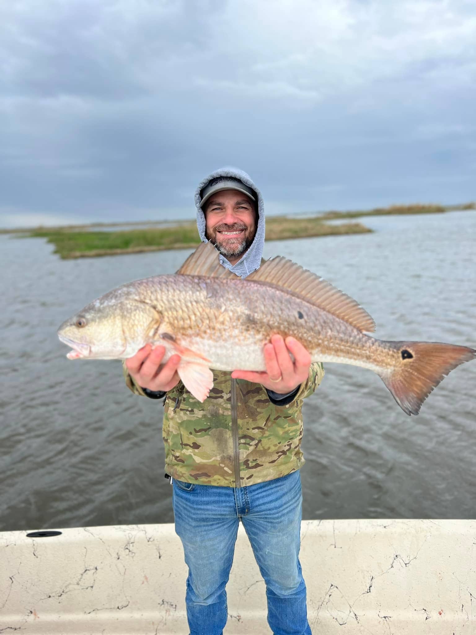 Man standing on a boat. he is holding a large bull red fish and smiling for the camera. The marsh, with its calm water, and green tall grass, is in the background. The sky is overcast.