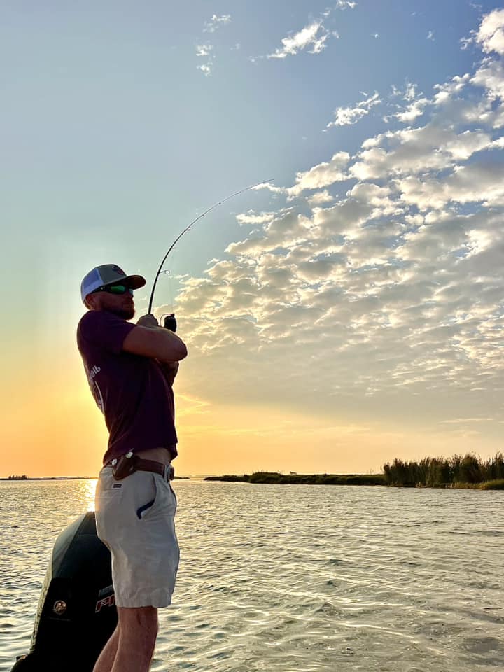 A man standing tall as he reels in a fish. He is on the back of a stationary boat. The water extend into the background and meets the yellow horizon, where the sun is rising.
