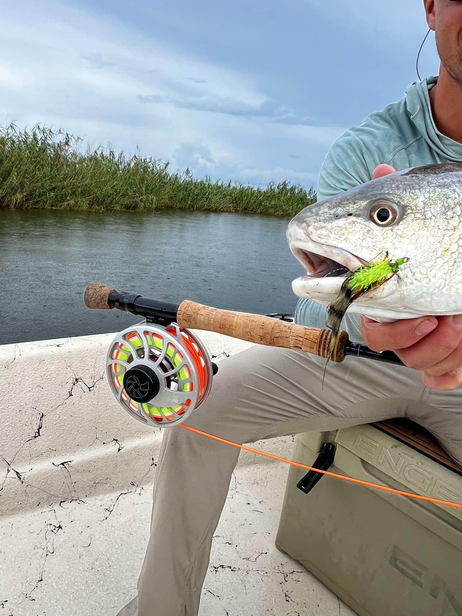 Up close photo of fisher showing off his catch. The fisherman is off to the right of the frame and his face is not visible. The fishes face is just visible in the right of the frame. In center frame is the bottom end of the fishing pole (complete with reel) resting on the man's knee. The left side of the frame captures the boat frame and the water and grass of the marsh.