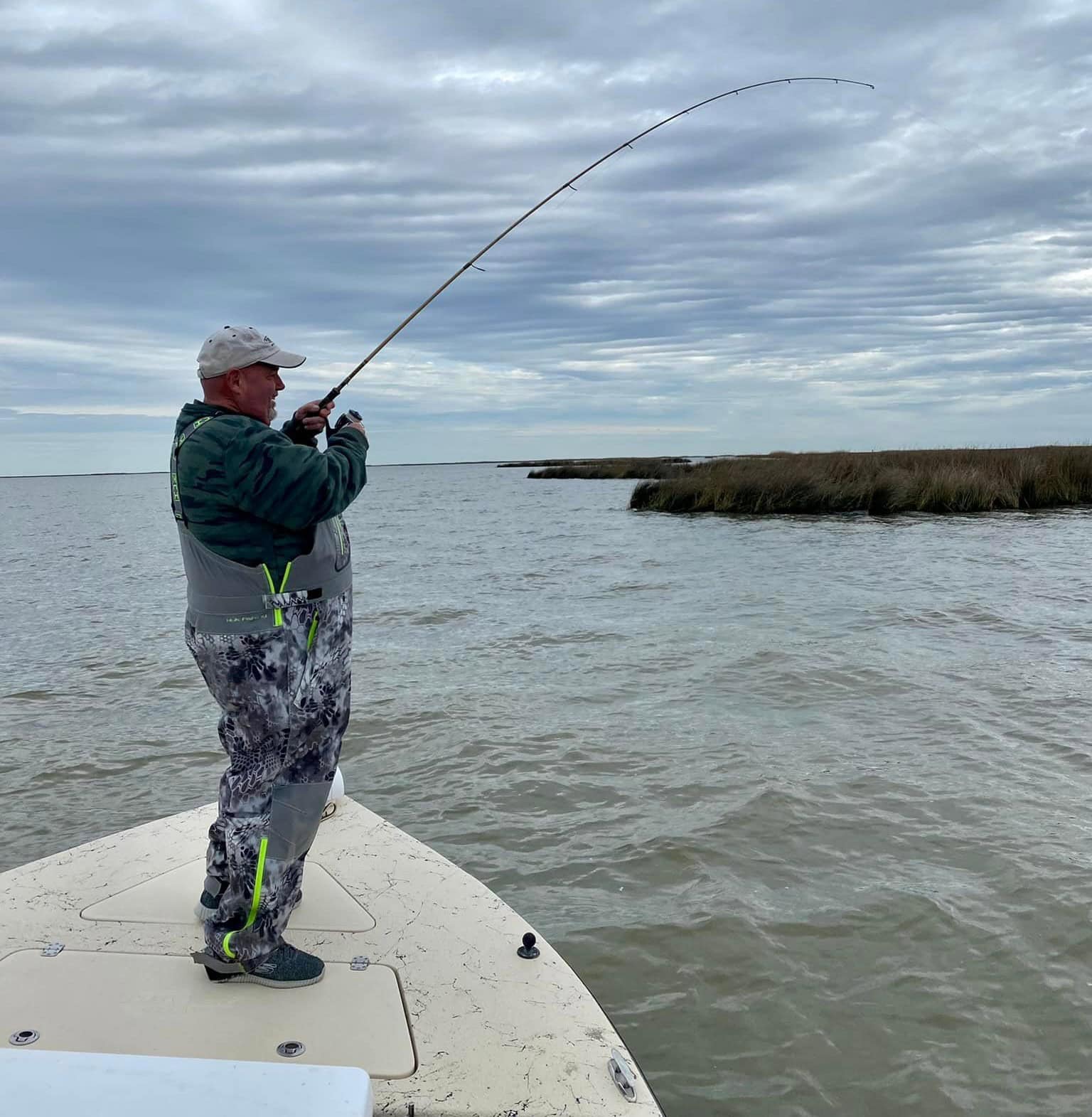 camo angler 2 Man in camouflage overalls on overcast day fishing from the bow of a boat. Marsh is in the background.