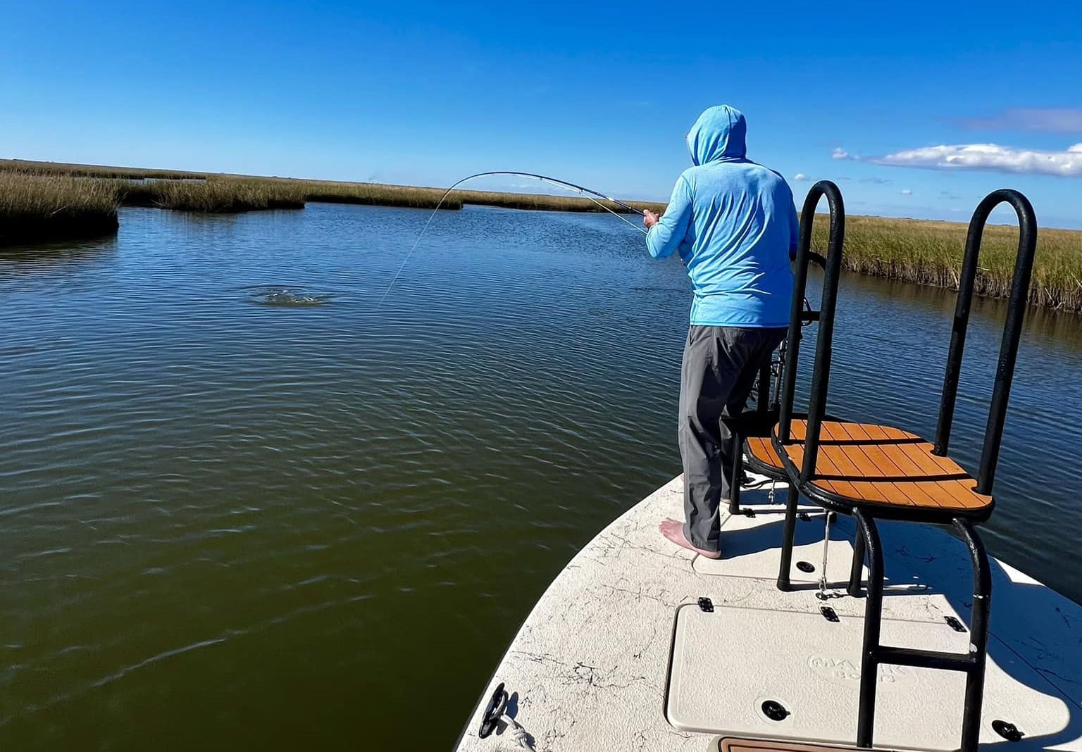 cool marsh angler shot 2 Man in blue hoodie fishing from the bow of a boat. Its a clear sunny day and the blue water and sky are broken up only by the tan reeds.