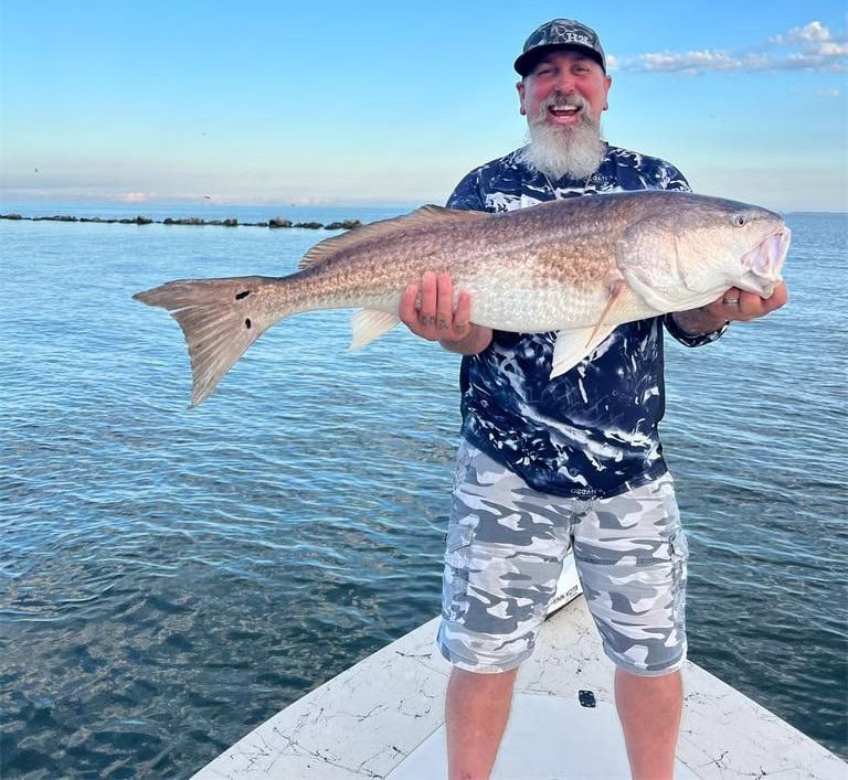 A smiling bearded man smiling from the prow of a boat as he holds up a large Redfish.
