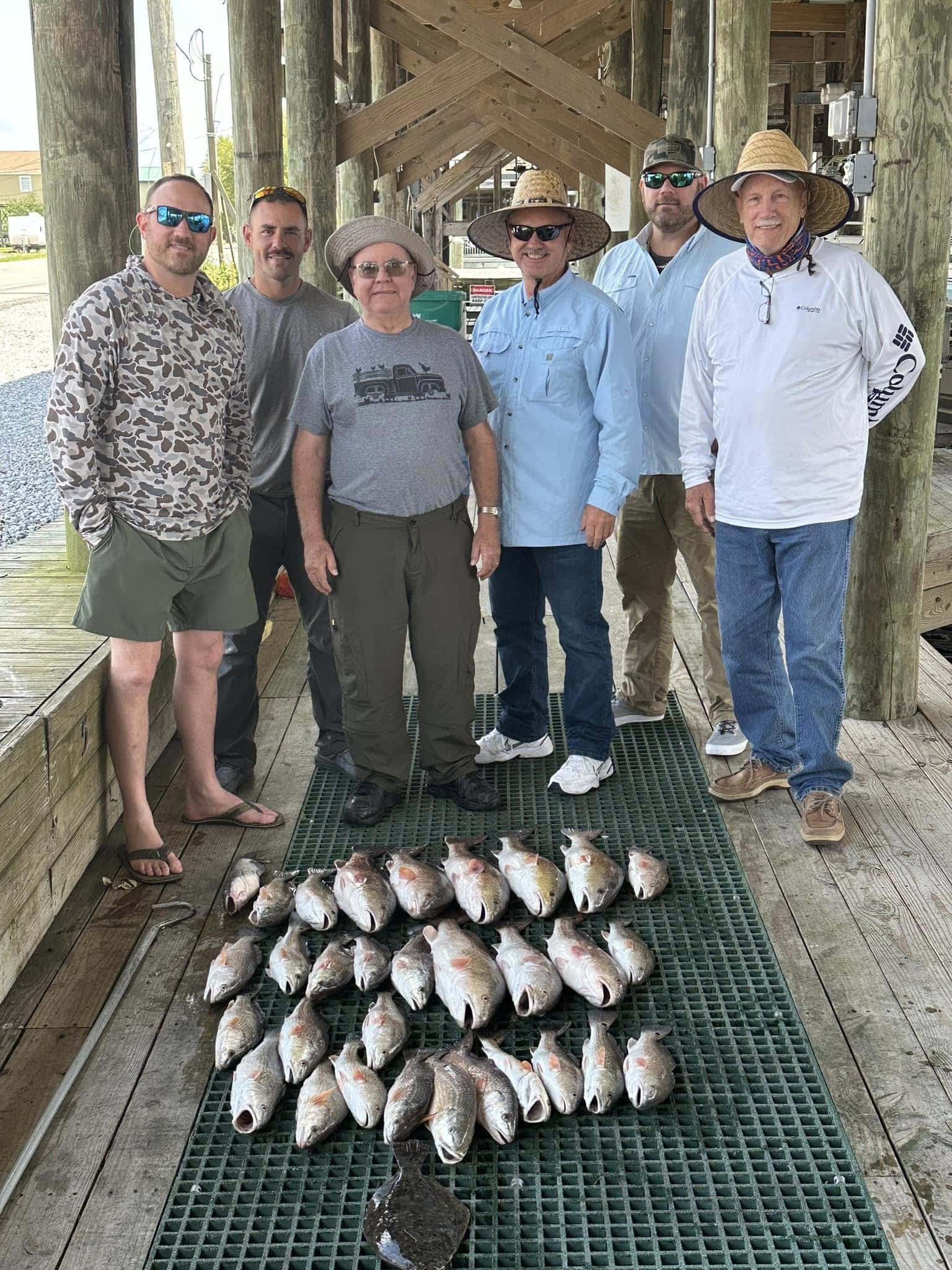 Six men standing beneath a pier and in front of their catch for the day which consists of about 27 fish.