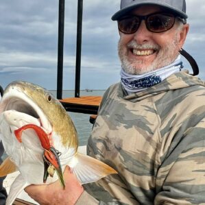 Man in hat and sunglasses showing off his catch to the camera. A red lure still hangs from the fish's mouth.