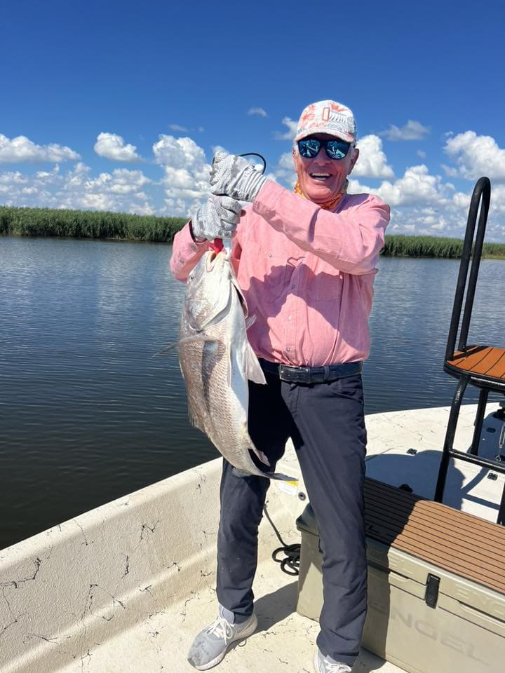 Man in a pink shirt standing in a boat holding up the fish he just caught.