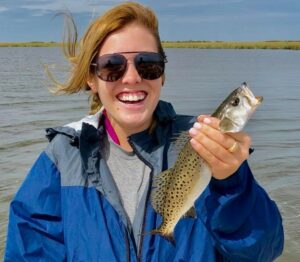 Lady wearing sunglasses and holding a speckled trout in her hand as she smiles for the camera Behind her are soft rippling waves of water.