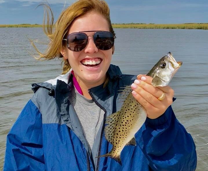 Lady wearing sunglasses and holding a speckled trout in her hand as she smiles for the camera Behind her are soft rippling waves of water.